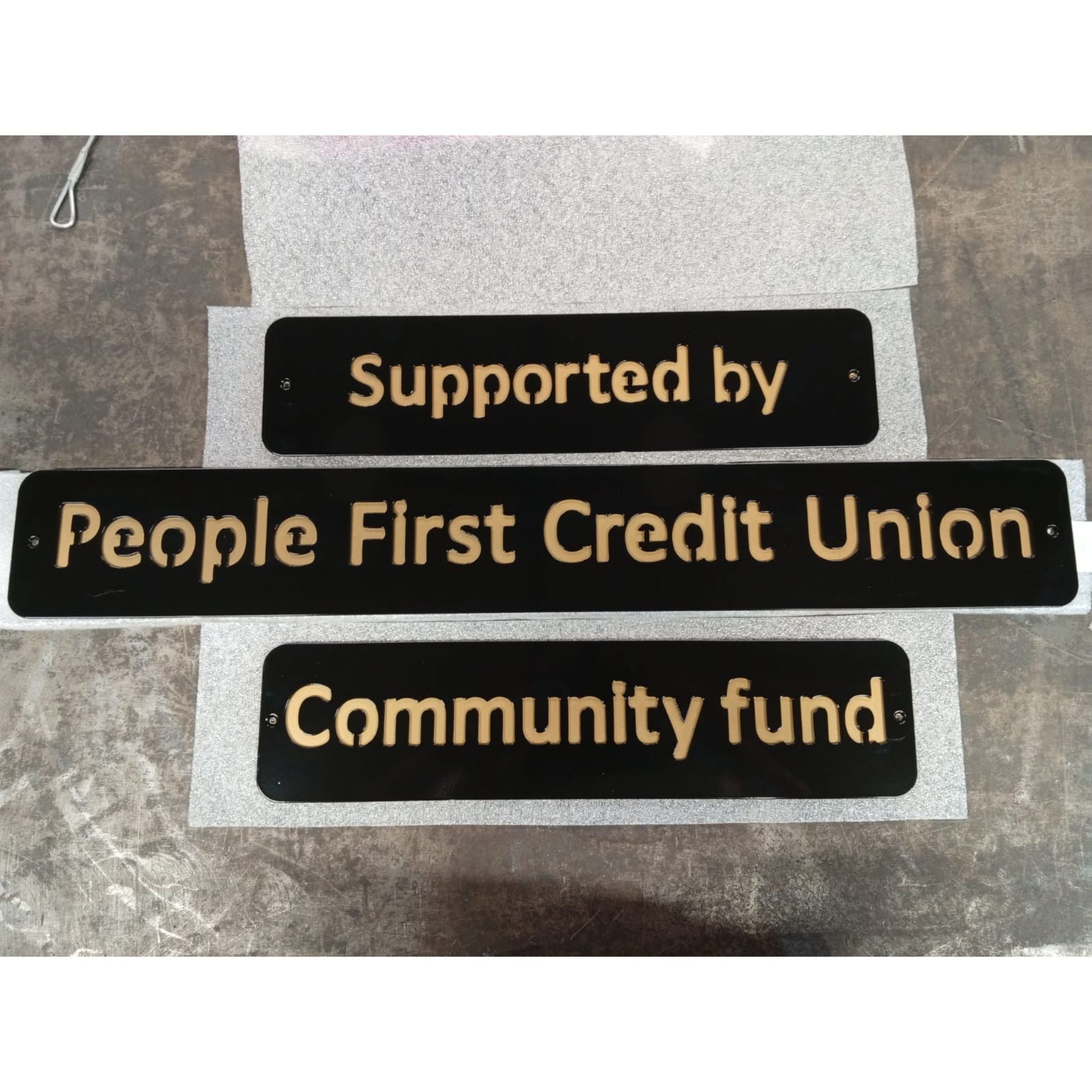 Three black signs with gold text on a gray background, supported by People First Credit Union Community Fund.